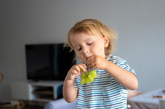 Photo little boy is sitting on a table in the kitchen and eating grapes. child is tasting healthy food.