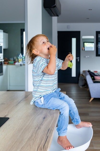 Little boy is sitting on a table in the kitchen and eating grapes. Child is tasting healthy food.