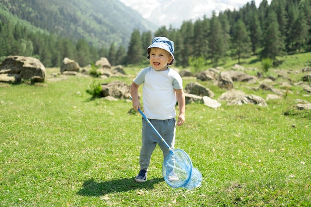 小さな男の子は、晴れた夏の日に蝶の網を持って歩いて、緑の丘で蝶を捕まえています