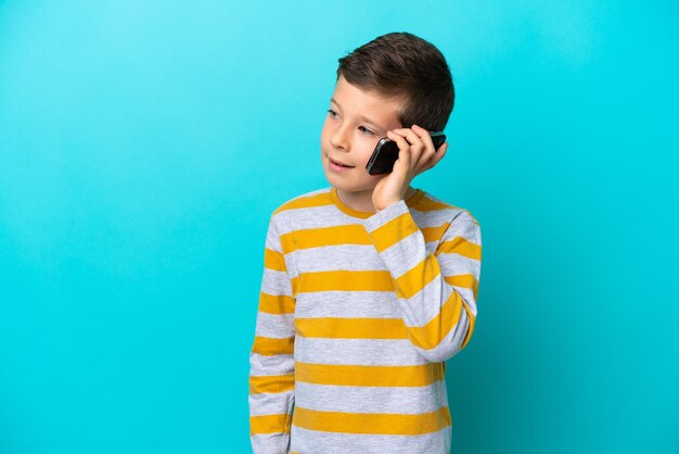 Little boy isolated on blue background keeping a conversation with the mobile phone with someone