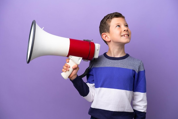 Little boy isolated on purple background holding a megaphone and looking up while smiling