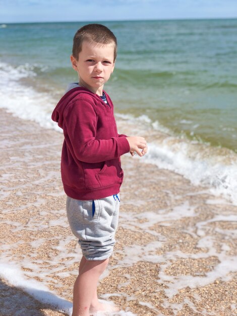 A little boy in a jacket and pants in sunny day stands on the seashore and looks to the camera. Recreation and tourism concept.