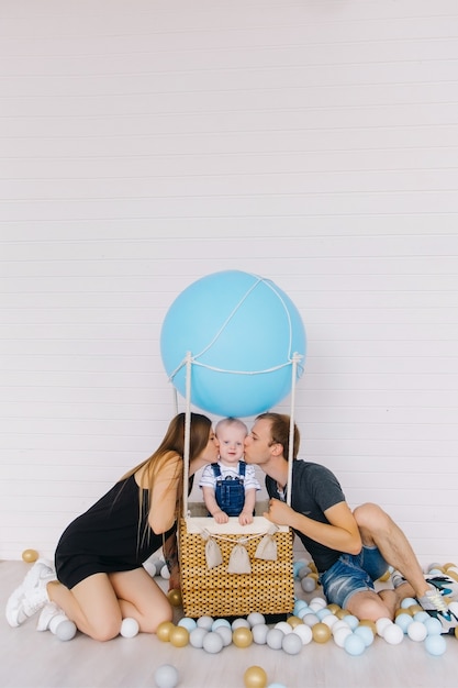 Little boy in jeans on blue balloon on white with his family. Parents are kissing they son.