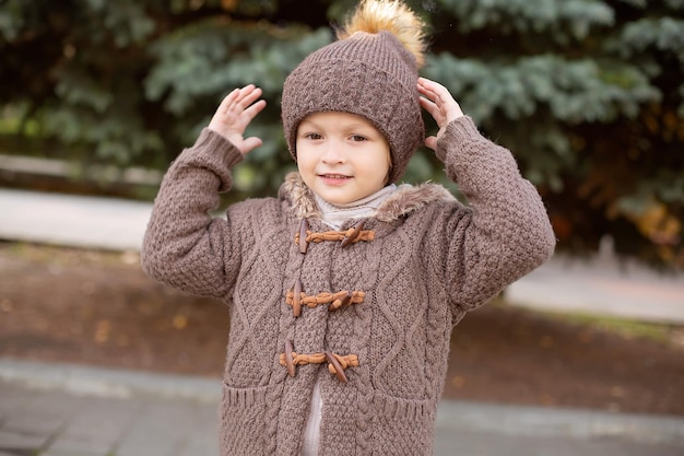 Photo a little boy in knitted things a hat and a brown jacket on the street touches a hat