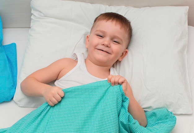 Little boy laughs as he lies in bed under ablue wool blanket. Happy morning.