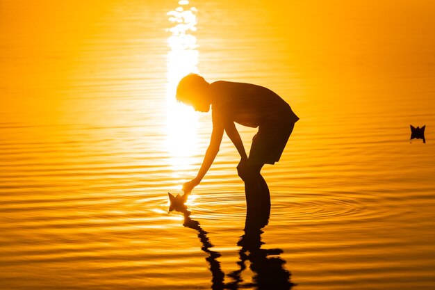 小さな男の子が夏にビーチで紙の船を打ち上げる美しい夕日折り紙川湖