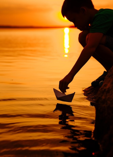 小さな男の子が水上で紙の船を打ち上げる美しい夏の日没紙の船折り紙
