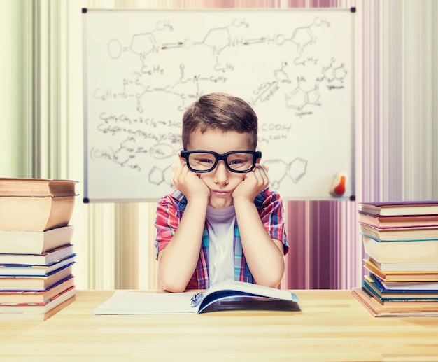 Маленький мальчик учится уроки в школьной библиотеке. Ученик в очках против книжных полок