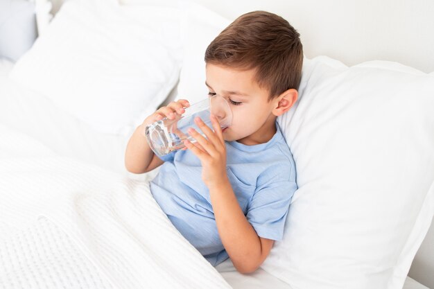 Little boy lies sick in white bed and drink water