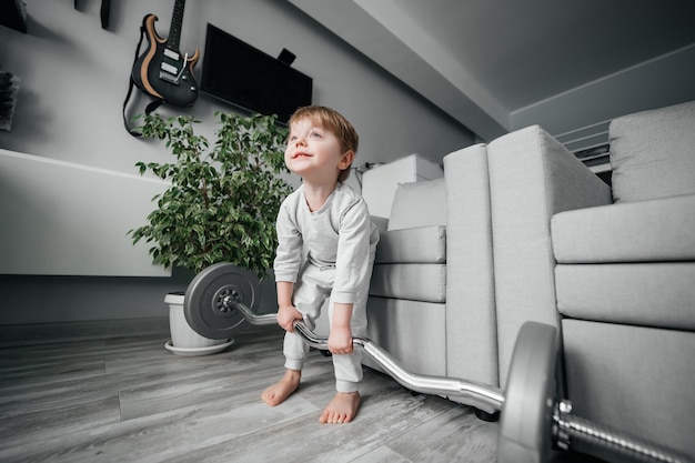 小さな男の子は灰色の背景の部屋でバーベルを持ち上げます