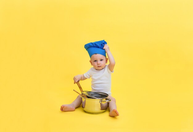 A little boy looks  and a chef's hat with a pot and a wooden spatula spoon. A boy in a white bodysuit pulls on a cap