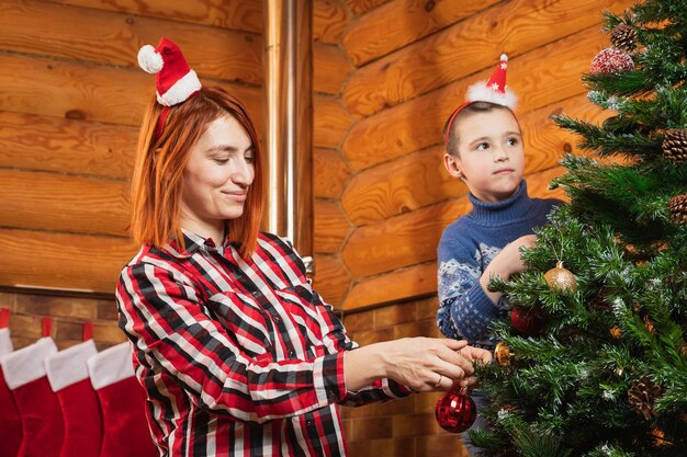 小さな男の子と母親がカントリーハウスで人工的なクリスマスツリーをガラス玉で飾ります