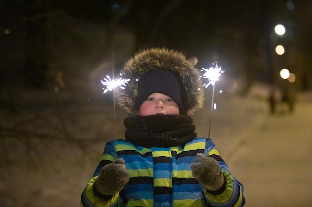 Маленький мальчик в ночном зимнем городском парке в теплом пальто с бенгальским огнем