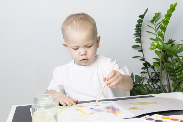 A little boy paints with watercolor paints on a table next to a green flower