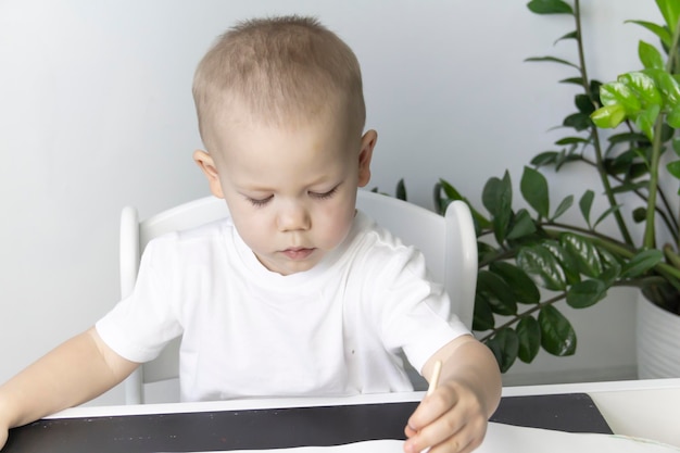 A little boy paints with watercolor paints on a table next to a green flower