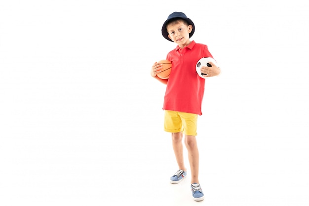 A little boy in panama, yellow jersey, red shorts and white sneakers stands with basketball and soccer balls