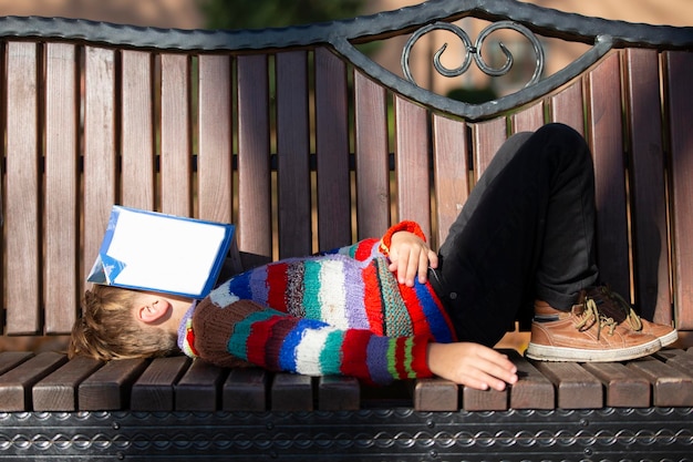 A little boy in the park lies on a bench and his face is covered with a book