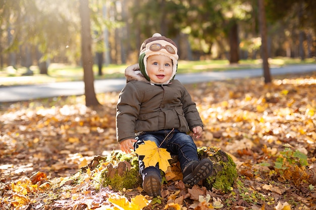 Little boy in the pilot cap sitting on the stump, yellow and orange foliage around him. Autumn