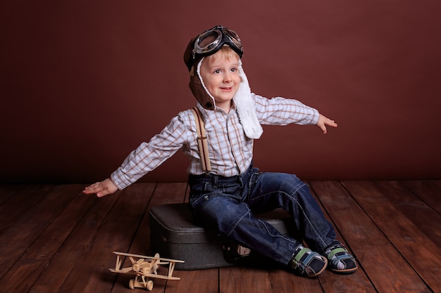 조종사의 헬멧을 쓴 어린 소년이 나무 비행기를 가지고 놀아요. 격자 무늬 셔츠와 멜빵에 소년.