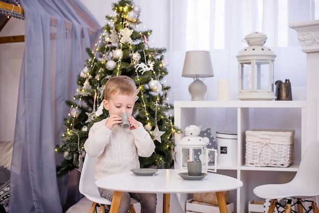 A little boy plays tea, drinks cocoa from a gray cup In the background there is a Christmas tree