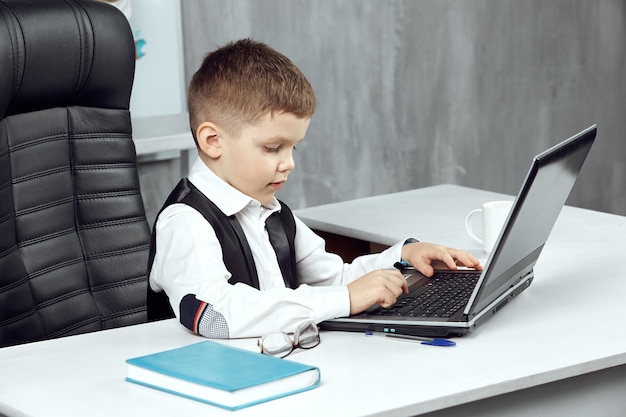 Маленький мальчик изображает босса в офисе, сидящего в кресле и работающего на ноутбуке