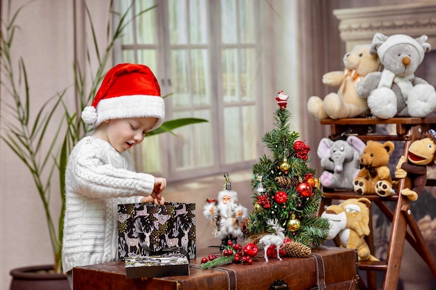 A little boy prepares gifts for Christmas and for his toys and decorates a small Christmas tree in a country house with a fireplace He is fun and interesting to play