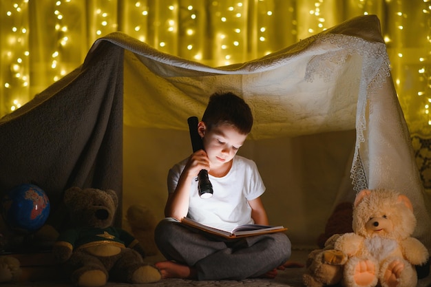 Little boy reading book in play tent at night