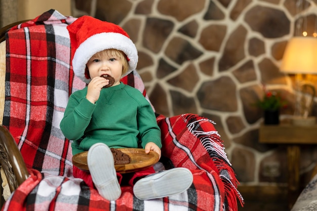 Little boy in red hat eating christmas gingerbread and chocolate chip cookies, waiting  santa claus at home for new year.