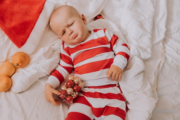 Little boy in red and white pajamas is lying in bed on a white sheet next to the Christmas tree. Christmas morning. healthy sleep of the child. lifestyle. space for text. High quality photo