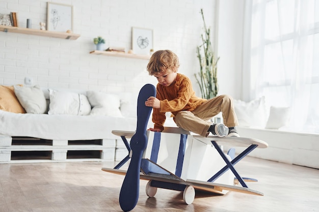 Little boy in retro pilot uniform having fun with toy plane indoors