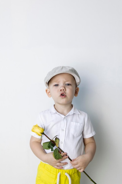 A little boy in retro style with a yellow rose in his hand stands near a white wall