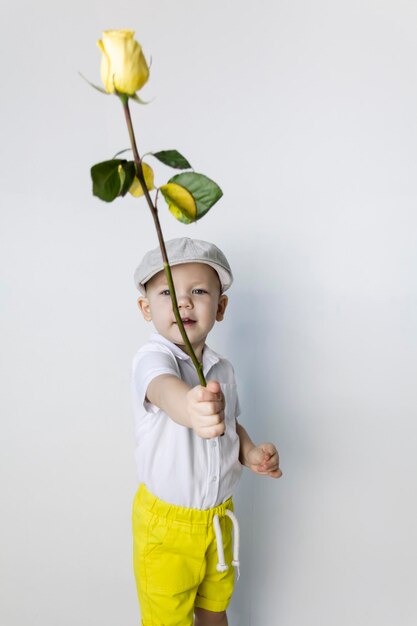 A little boy in retro style with a yellow rose in his hand stands near a white wall