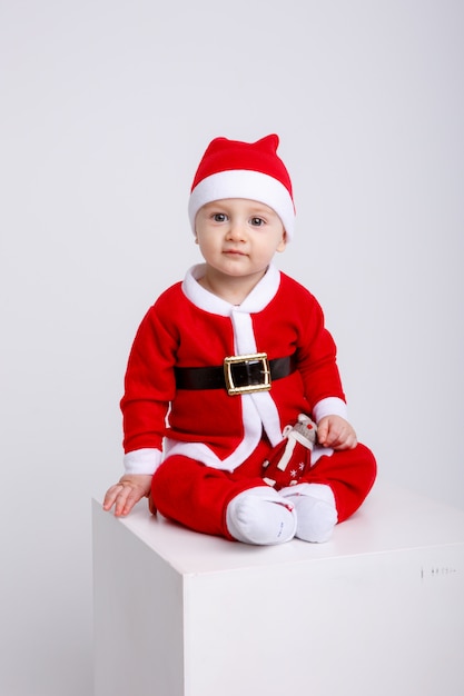 Little boy in Santa costume sitting on white cube on white background