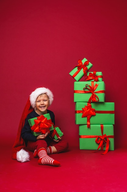 Little boy in Santa hat sits next to pyramid Christmas gift boxes on red in Studio.