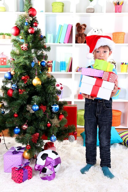 Little boy in Santa hat stands near Christmas tree with gifts