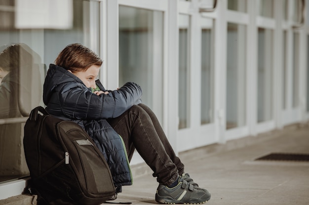 Little boy or school child sitting alone on floor in front of the school after suffering an act of bullying