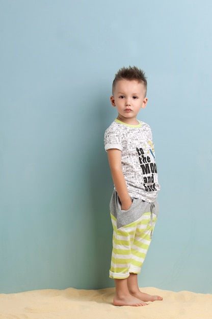 Little boy in a shirt and shorts standing on the sand on a light blue background