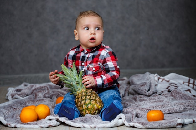 The little boy in the shirt sits on the plaid and holds the fruit