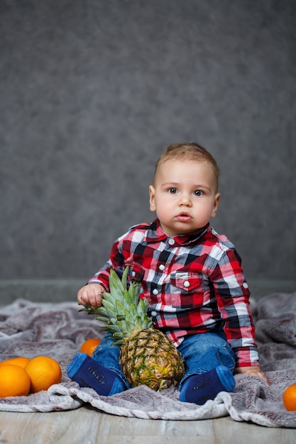 The little boy in the shirt sits on the plaid and holds the fruit