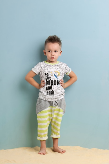 Little boy in shorts and t-shirt standing on the sand on a light blue background