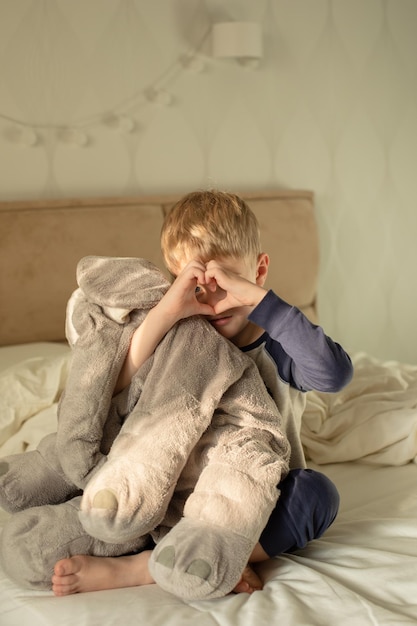 A little boy shows a heart with his hands sitting on the bed in the morning