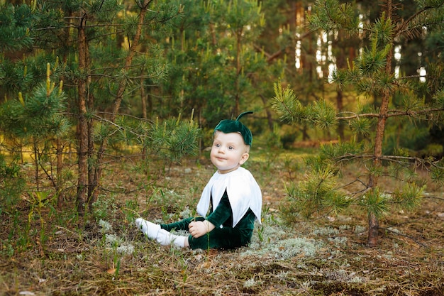 Photo little boy sits on a forest path in a snowdrop costume. boys dressed in a funny snowdrop costume with a hat