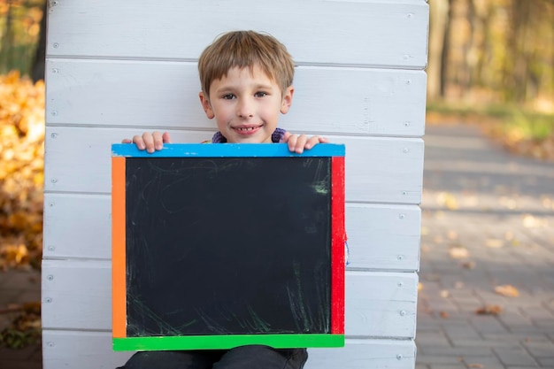 A little boy sits in the park near the wall and holds an empty black board for the inscription