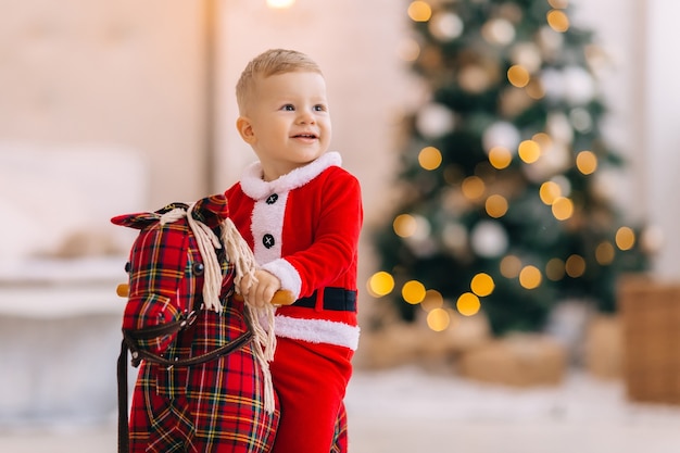 小さな男の子は木馬に座って微笑んでいます。ぼやけたクリスマスツリー