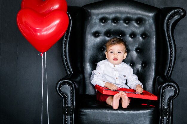 Little boy sitting on the armchair with red framed picture on the St. Valentine's day. 