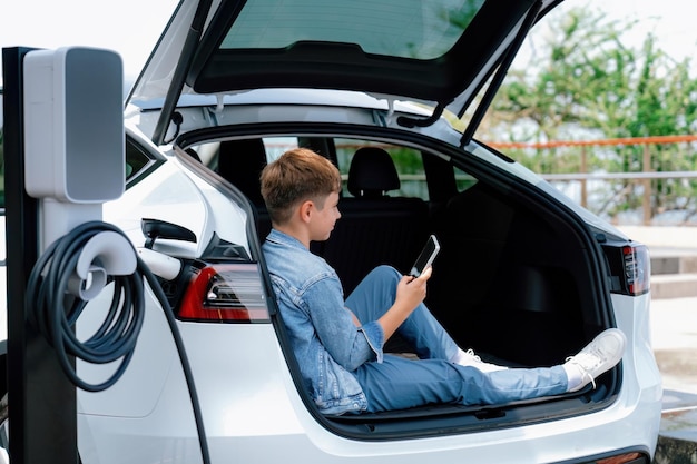 Photo little boy sitting on car trunk using smartphone while recharging ecofriendly car from ev charging station ev car road trip travel as alternative vehicle using sustainable energy concept perpetual