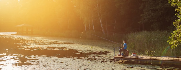 Little boy sitting and fishing on a wooden dock at the sunset