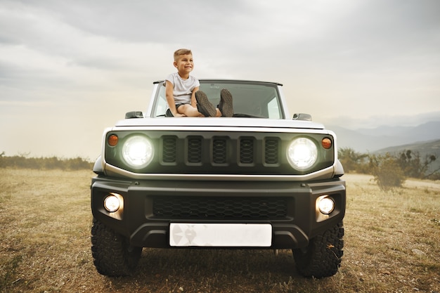 Little boy sitting on a hood of an offroad car parked in mountains