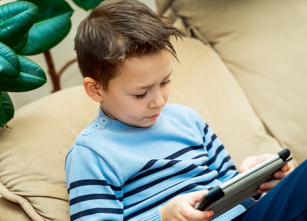 Little boy sitting on light couch and touching the screen of wireless tablet at home Portrait of a young child with a laptop