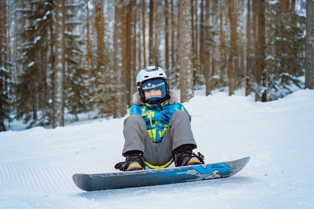 Little boy sitting on snow putting his feet in snowboard bindings adjusting straps